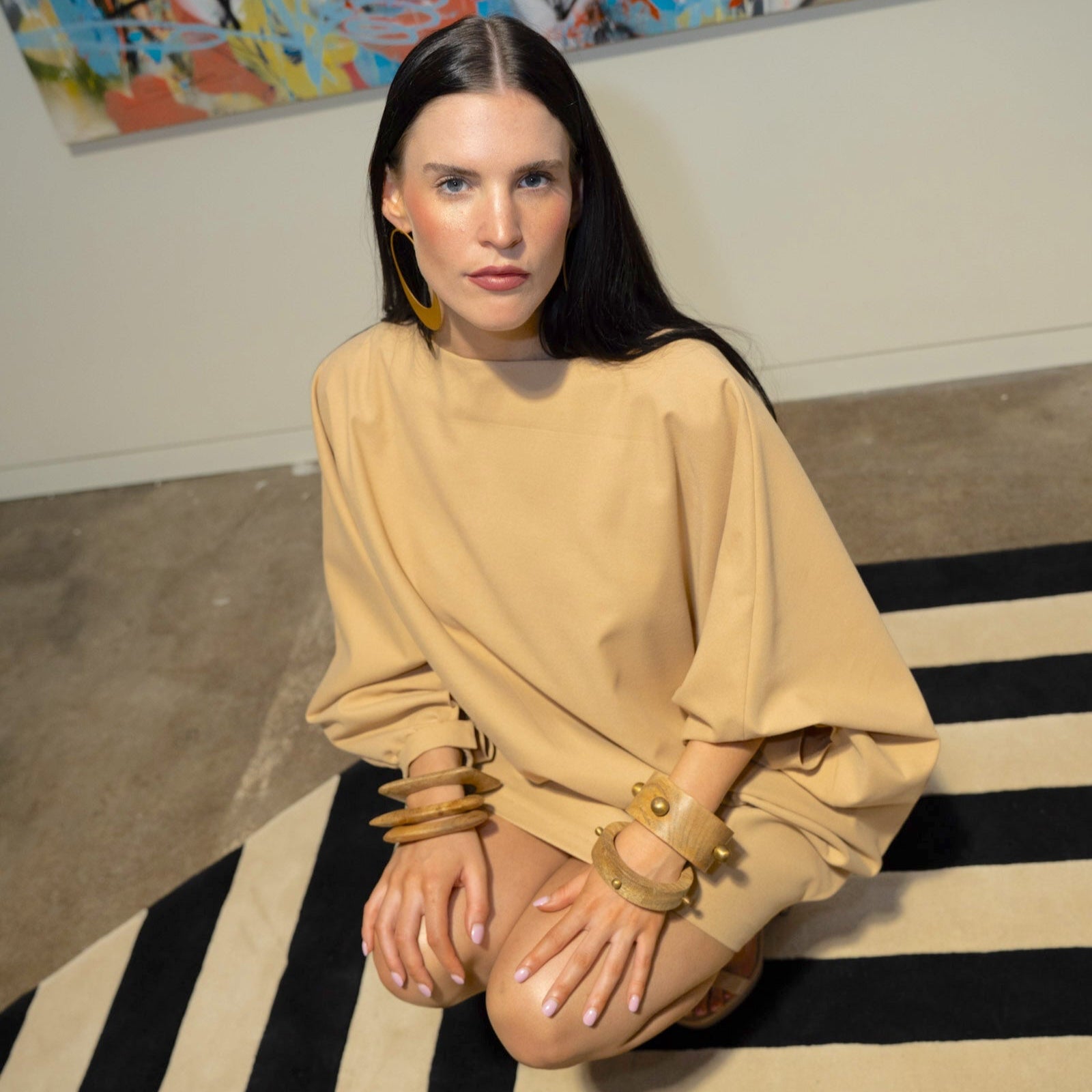 Woman in beige outfit sitting on a black and white striped rug with colorful abstract art in the background.