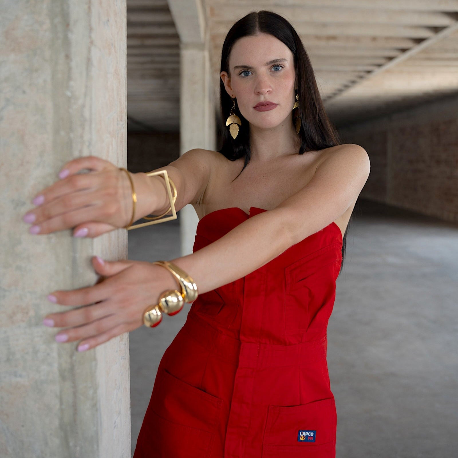 Woman in a red dress with gold jewelry standing against a stone wall.