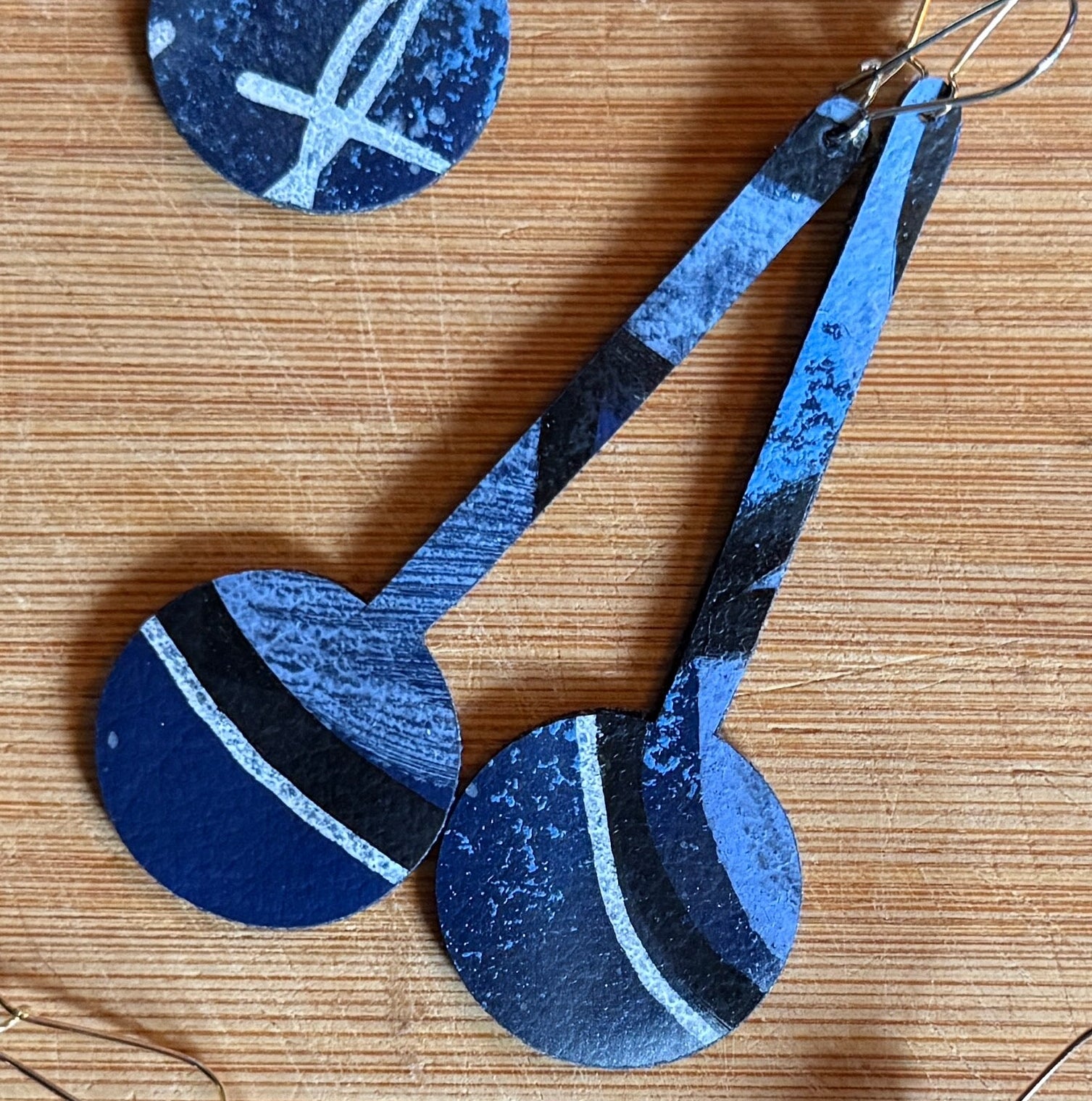 A collection of handmade earrings with various geometric and abstract patterns in shades of blue, red, yellow, and gray, displayed on a light wood background.