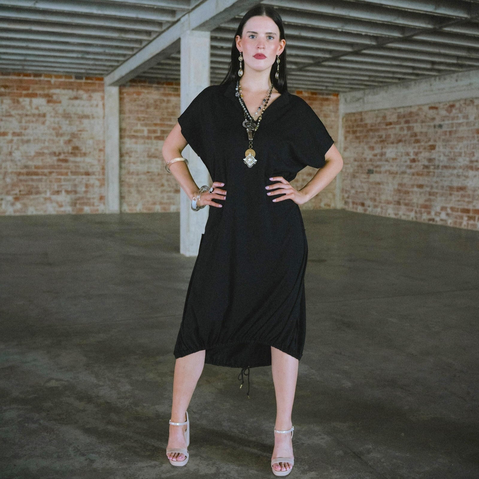 Woman in a black dress standing in an industrial-style room with brick walls and concrete floor.