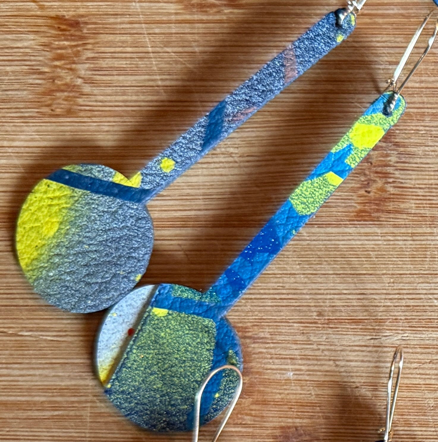 A set of handmade dangle earrings with circular disc charms and patterned rectangular drops, displayed on a wooden surface. The earrings feature various colorful abstract patterns in shades of blue, red, yellow, and gray.