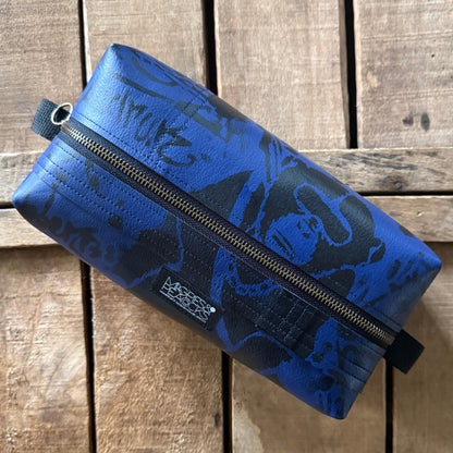 A blue zippered pouch with black graphic graffiti featuring a small Magpies & Peacocks logo patch, resting on a rustic wooden plank background.