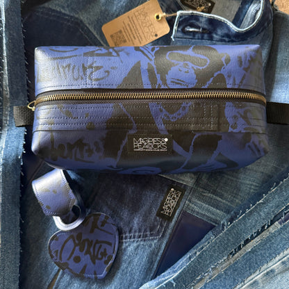 A blue and gray graffiti-patterned zippered pouch from Magpies & Peacocks styled on top of a denim jacket on a wooden surface.