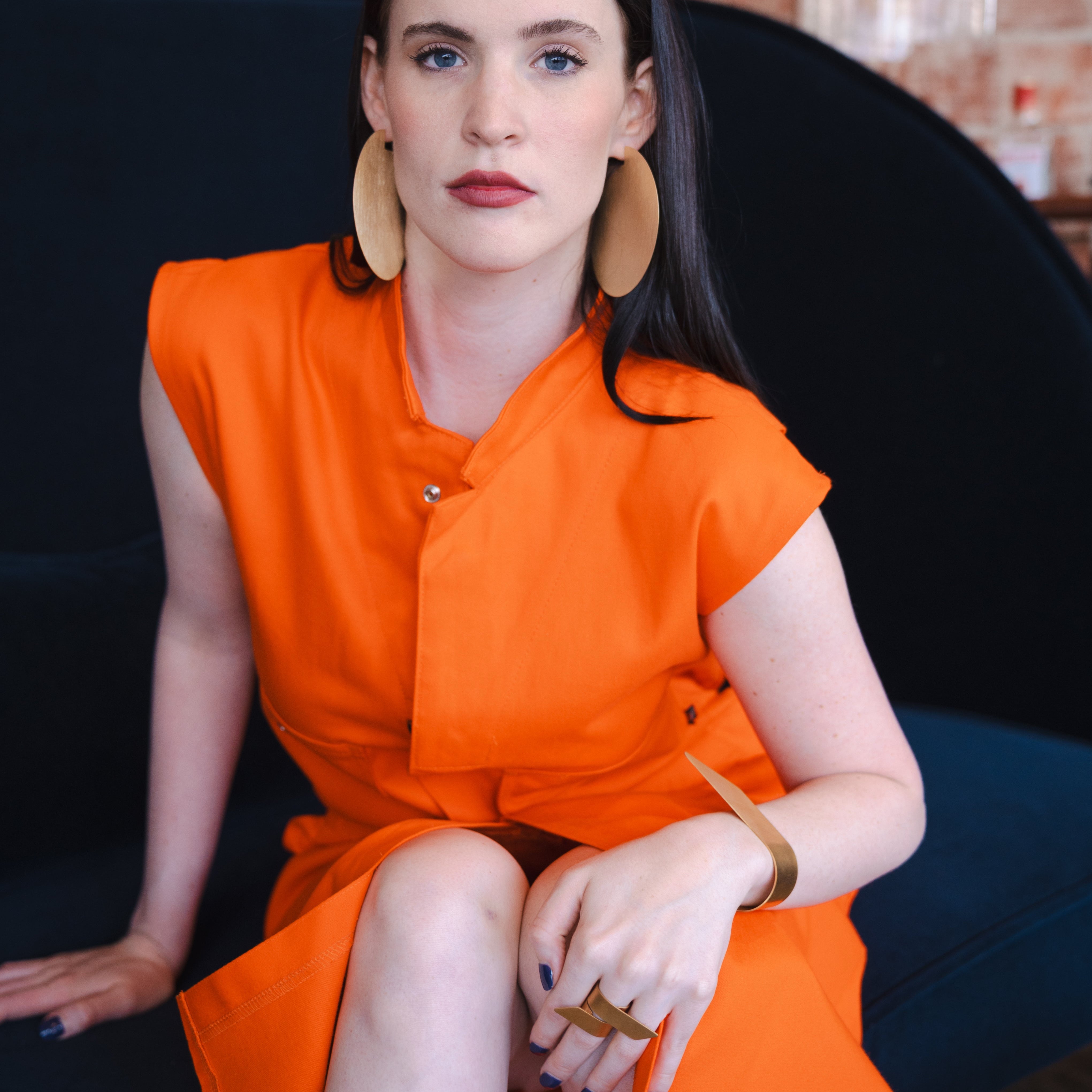 Woman in an orange dress sitting on a black chair with a brick wall background