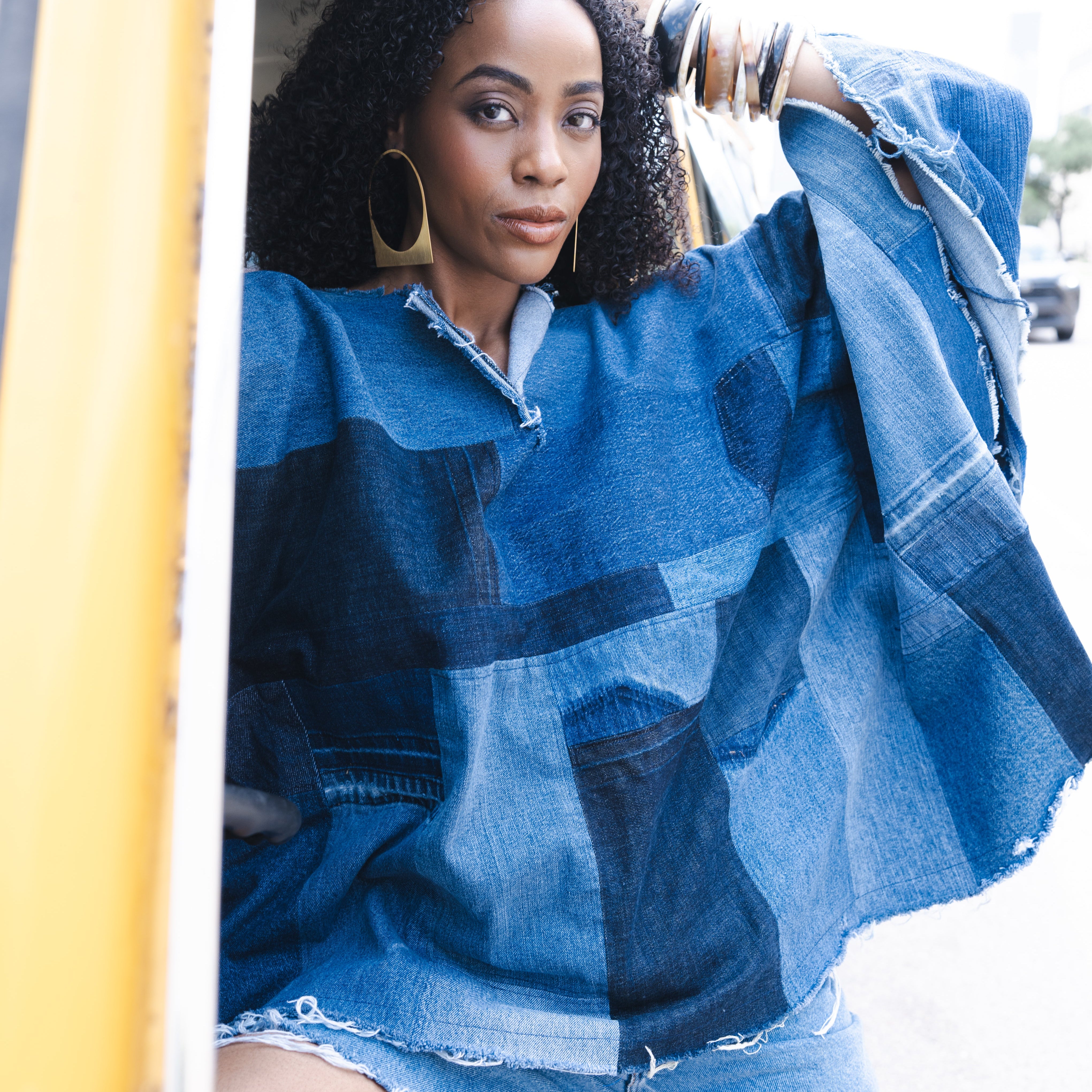 Woman wearing a blue upcycled denim poncho and shorts sitting inside a yellow vehicle.