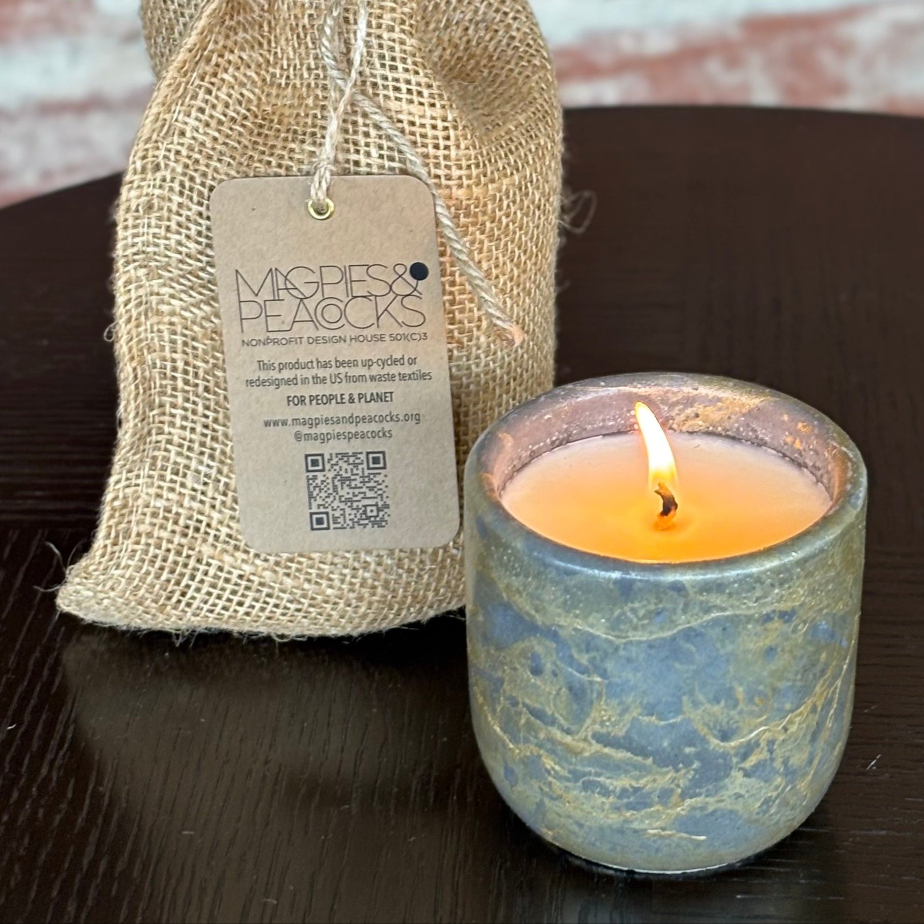 Candle in a decorative holder next to a burlap bag with a tag on a wooden surface.