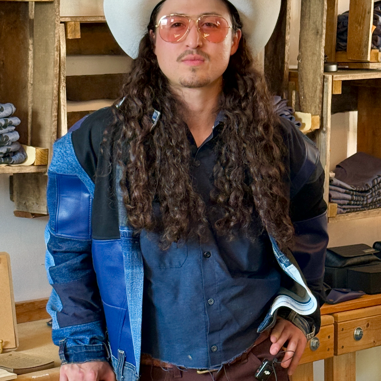 Person wearing a cowboy hat and sunglasses in a rustic setting with wooden shelves.