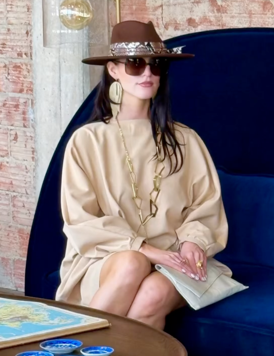 Woman sitting on a blue couch wearing a beige dress, brown hat, and sunglasses in a room with a brick wall.