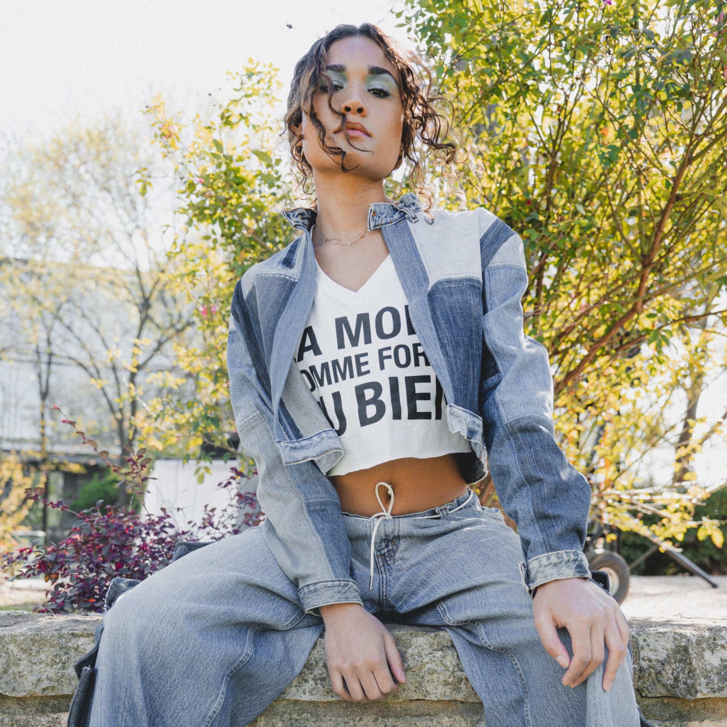 A person sitting on a stone bench wearing a crop top paired with a pieced denim jacket and blue jeans.